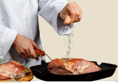 A chef sprinkles sea salt over a large piece of meat in a skillet.
