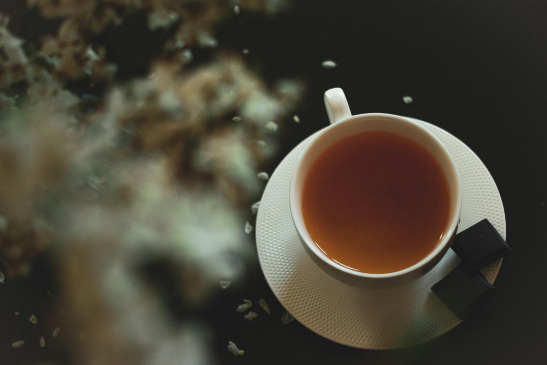 An image of a porcelain cup on a saucer with tea and blurred herbs in the foreground.
