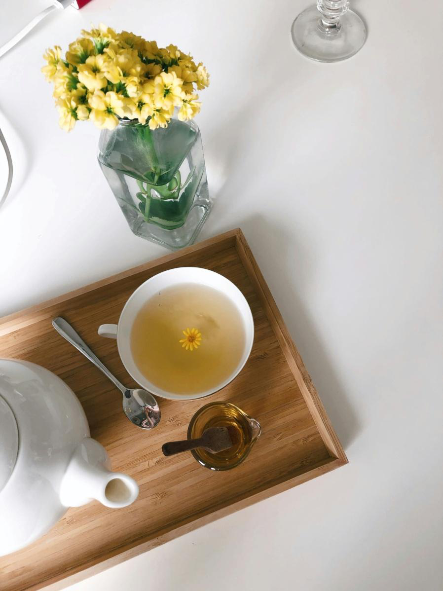 A warm cup of herbal tea on a wooden tray with honey and a small yellow flower floating on top beside a vase of yellow blossoms.