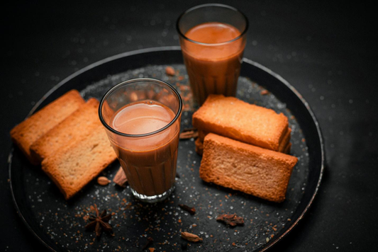 An aesthetic image of a tray with two cups of Indian tea, scattered spices and rusk.