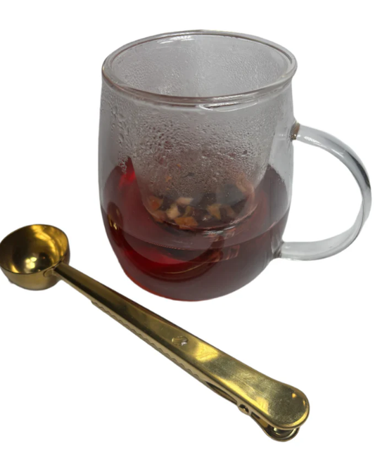 Glass mug of deep red tea gently brewing with a gold measuring spoon beside it, highlighting chef-style brewing precision.