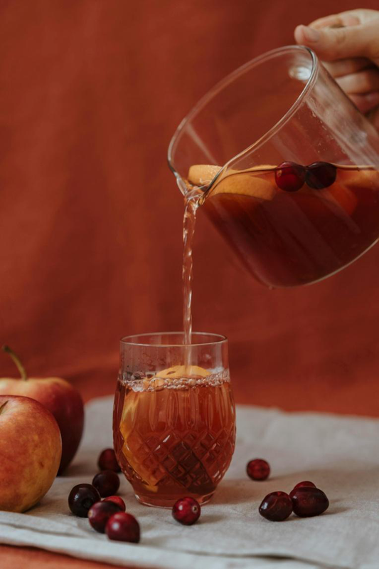 A glass pitcher pouring cranberry and apple infused fruit tea into a clear glass, surrounded by fresh apples and cranberries.