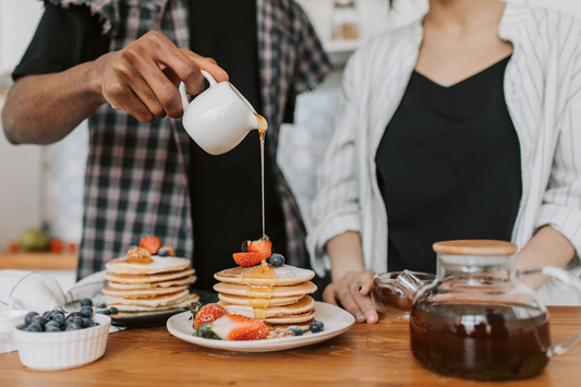 A person pouring syrup over a stack of fluffy pancakes, surrounded by a teapot, cup, and blueberries.