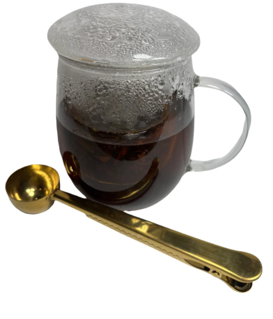 A glass mug of freshly brewed tea with a golden measuring spoon beside it.
