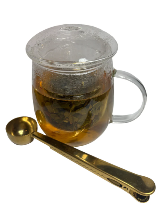Glass tea cup with lid and golden tea spoon beside it.
