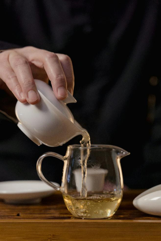 A hand holding a ceramic gaiwan, pouring fresh green tea into a glass teapot.