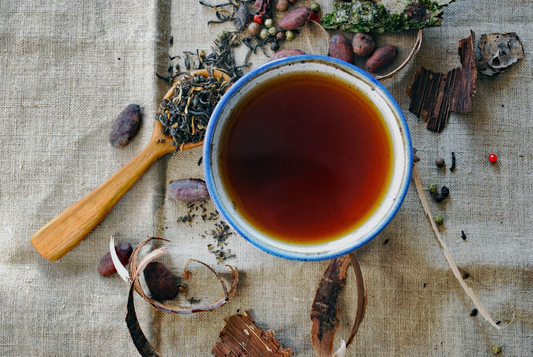 Overhead view of brewed rooibos tea in a ceramic cup surrounded by loose tea leaves, spices, and natural ingredients on linen fabric