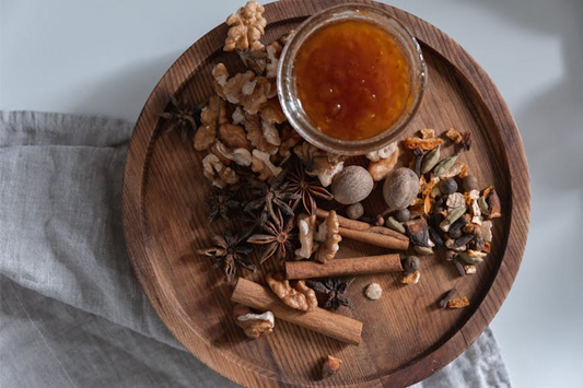A wooden plate with a glass jar of honey surrounded by assorted spices