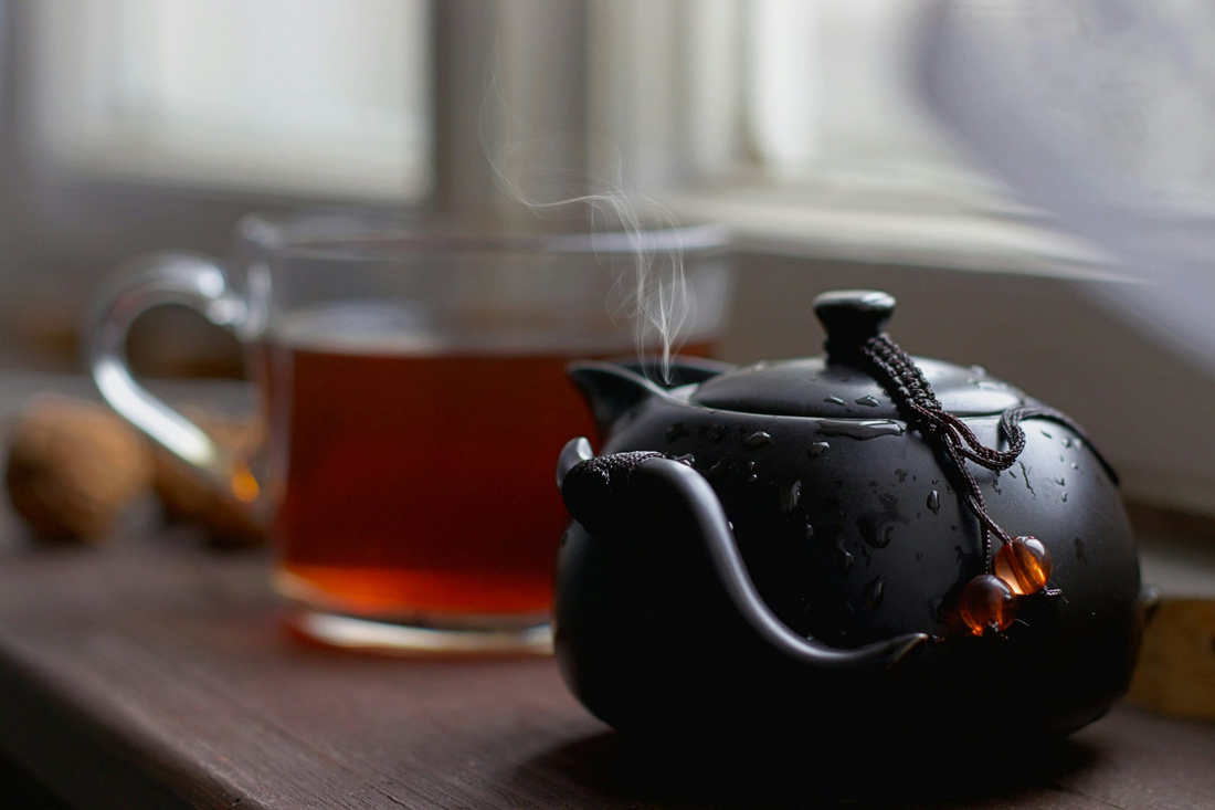 An image focused on a black teapot with steam rising from its nozzle and a glass cup filled with tea in the background.